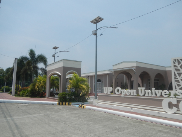 Offener Universitätscampus-Eingang mit einer Straße, die zu einem Gebäude führt, umgeben von Bäumen und Pflanzen, eine texttragende Wand auf der rechten Seite und sichtbarer Himmel im Hintergrund.