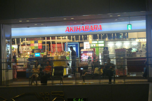 Ein Geschäft mit zwei Bänken davor, auf denen jeweils einige Personen sitzen, einem Schild mit Text darüber und einer Zaunwand unten.