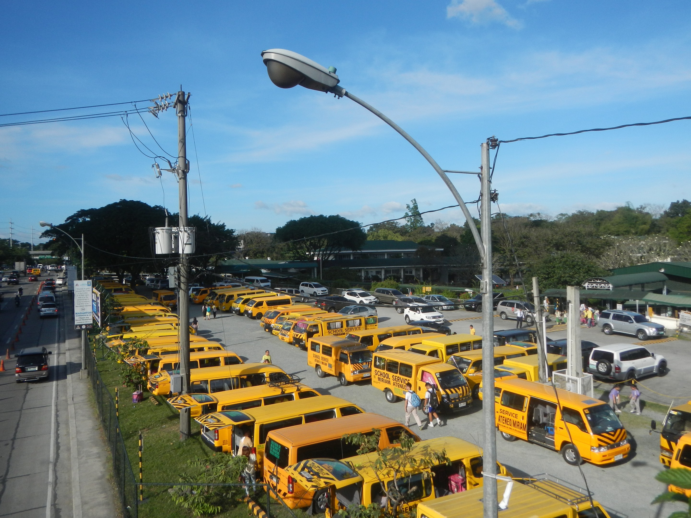 Eine Reihe gelber Schulbusse, die entlang einer Straße geparkt sind, mit Fußgängern auf dem Gehweg, Strommasten, Bäumen, Gebäuden und einem bewölkten Himmel im Hintergrund.
