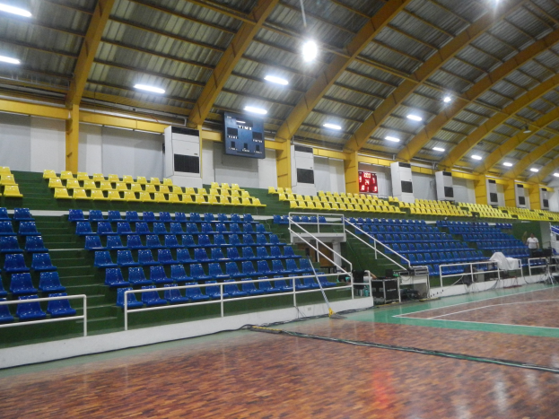 Groes Indoor-Sportstadion mit Holzfussboden, blauen und gelben Sitzpltzen, Gelndern, Treppen mit Gelndern, wandmontierten Bildschirmen, Deckenleuchten und einigen Personen.