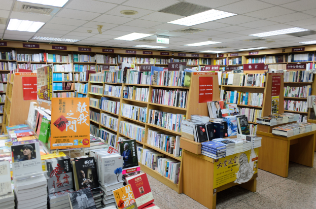 Ein gut organisierter Buchladen mit ordentlich aufgestellten Büchern auf Regalen und Tischen, beleuchtet von Deckenlampen und mit informativen Schildern auf dem Boden.