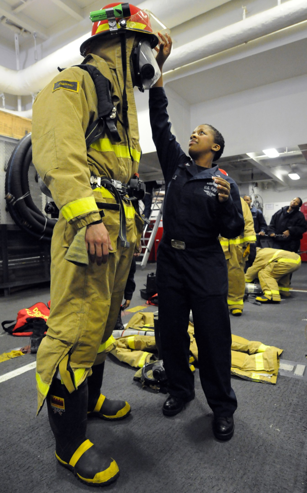Zwei Feuerwehrleute, einer in einem schwarzen Kleid und Helm und ein anderer in gelb-schwarzer Ausrüstung, stehen in der Nähe einer Leiter und anderer Ausrüstung mit Menschen im Hintergrund.