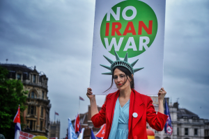 Eine Frau steht in der Mitte und hält ein Schild mit der Aufschrift "Kein Iran-Krieg" mit einer Freiheitsstatue auf dem Kopf, mit Menschen, Flaggen, Gebäuden, Bäumen und einem klaren blauen Himmel im Hintergrund.