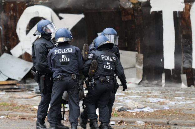 Polizisten in schwarzen Uniformen und blauen Helmen stehen vor einem mit Graffiti bedeckten Gebäude während einer Ausschreitung, mit verstreuten Papieren und Gegenständen auf dem Boden.