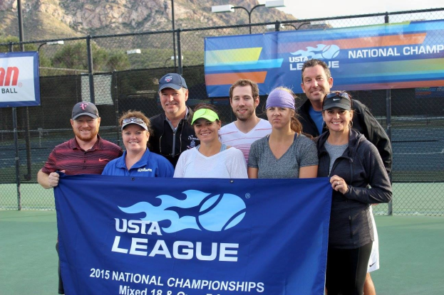 Gruppe von Menschen auf einem Tennisplatz mit einer "2015 USTA National Championships"-Fahne, einem Zaun, Fahnen, Laternenmasten, Bäumen, Bergen und einem klaren blauen Himmel im Hintergrund.