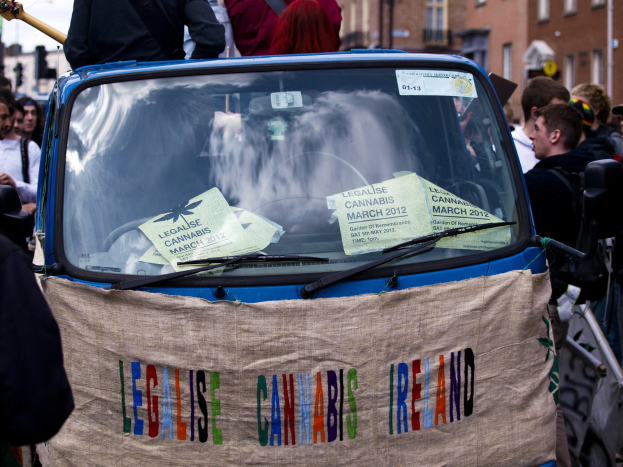 Gruppe von Menschen um ein Auto mit einem "Legalise Cannabis Ireland"-Schild, das Papier im Fahrzeug hält, mit Gebäuden, Laternen und einem klaren blauen Himmel im Hintergrund.