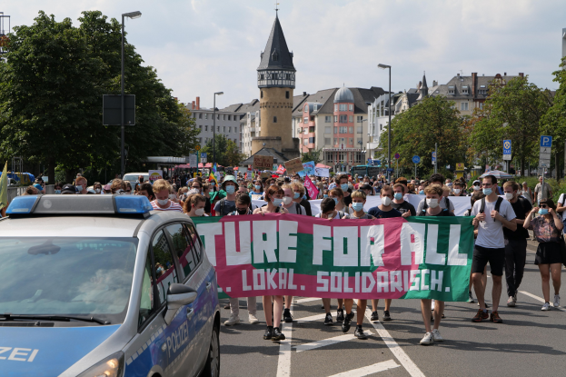 Eine große Gruppe von Menschen marschiert mit einem "Lokal Solidarität"-Schild die Straße entlang, mit einem Polizeiwagen auf der Seite, Bäume und Gebäude sind im Hintergrund unter einem bewölkten Himmel zu sehen.