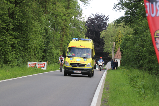 Ambulanz fährt auf einer Straße mit Fahrradfahrern daneben, gesäumt von Gras, Bäumen, Häusern, Strommästen und einem klaren blauen Himmel.