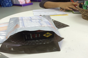 Ein Mann sitzt an einem Tisch mit einer Paketbox, einem Korb mit Einwickelpapier und einem Handy, mit mehreren Kisten hinter ihm.