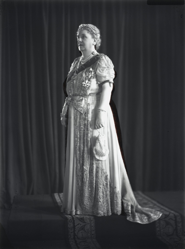 Schwarzes und weißes Foto von Queen Elizabeth II auf einer Bühne mit einem Vorhanghintergrund, die eine detaillierte königliche Robe und einen klassischen Hairstyle trägt und zur Seite schaut.