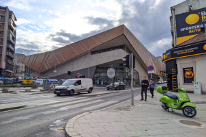 Ein grüner E-Scooter steht auf dem Straßenrand neben einem Gebäude, mit Fahrzeugen auf der Straße, Menschen auf dem Gehweg und Verkehrsampeln mit Masten, mit Gebäuden, Bäumen und einem bewölkten Himmel im Hintergrund.