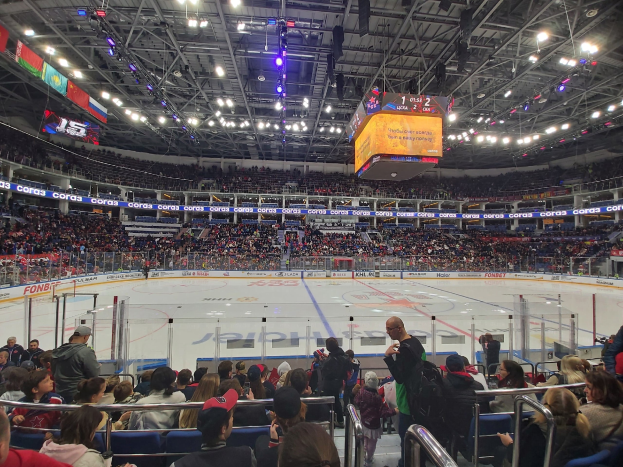 Eine große Menschenmenge in einer Hockeyarena, einige sitzen und einige stehen, schaut ein Spiel auf dem Eis, das von einem Zaun umgeben ist, mit hellen Lichtern und Bannern, die von der Decke hängen.