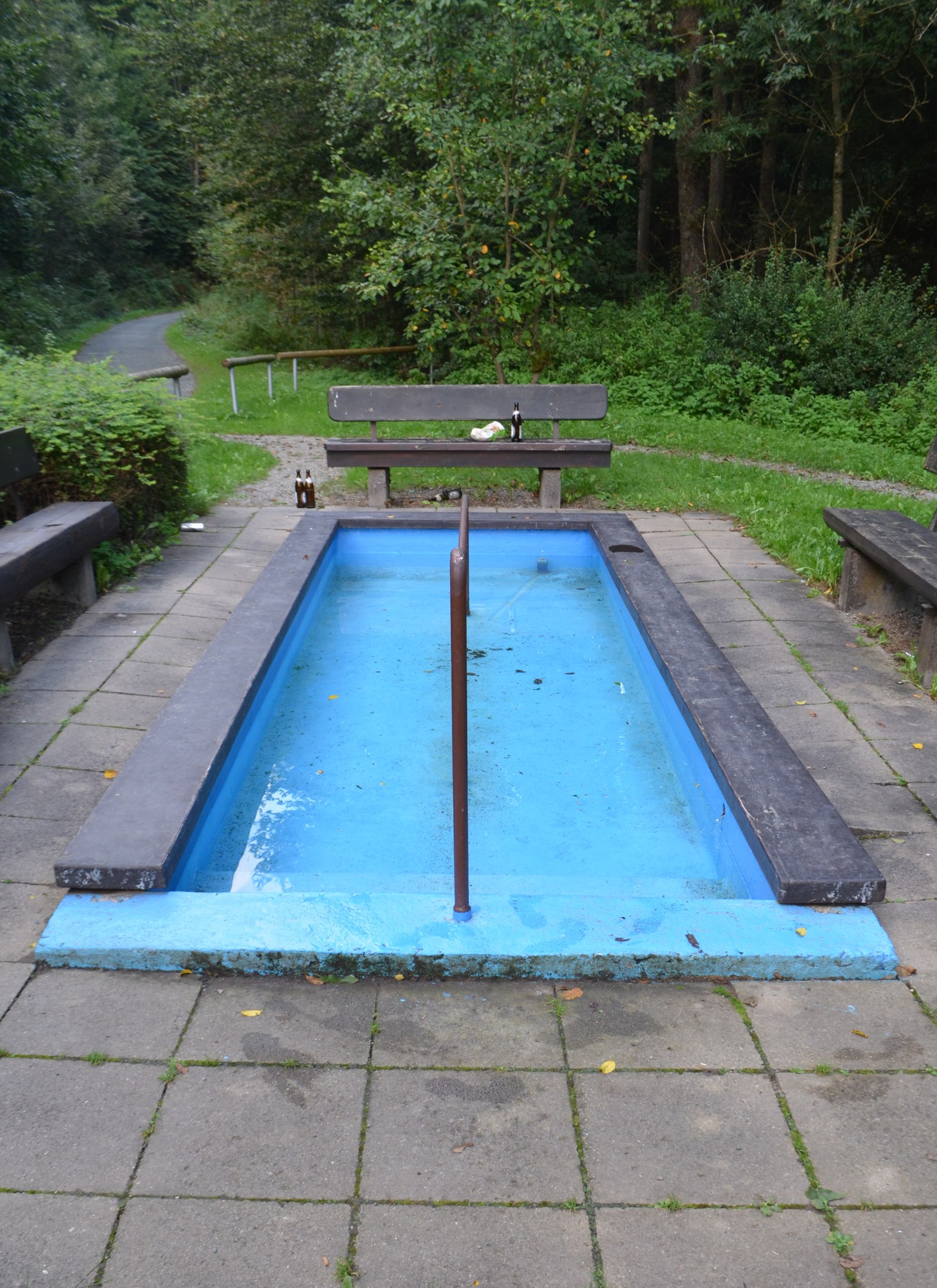 Kleiner Parkpool mit zentralem Pfahl, umgeben von Bänken, Grünflächen, Straße und Bäumen.