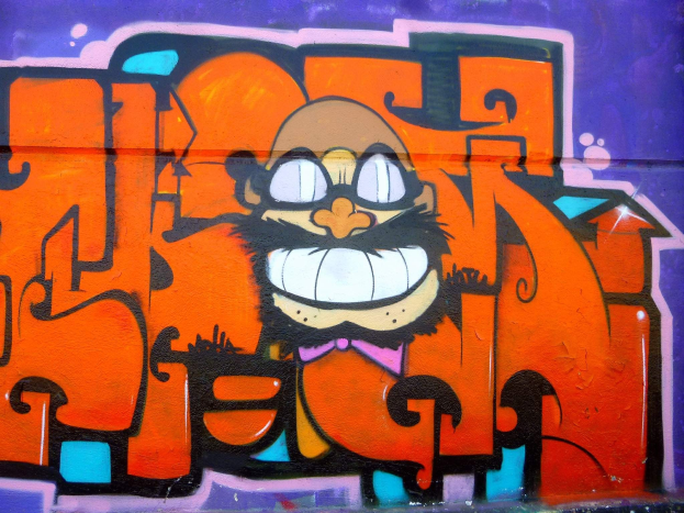 Graffiti-Gemälde eines Cartoon-Charakters mit großen Augen, einem breiten Grinsen und einem verschmitzten Ausdruck an der Seite eines Gebäudes, mit leuchtenden Farben und auffälligem Begleittext.