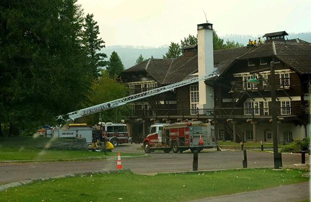 Feuerwehrfahrzeug vor einem großen Gebäude mit Fenstern, Geländern, Säulen und einem Schornstein geparkt, umgeben von Absperrbaken, Gras, Bäumen und Laternen, mit ein paar Menschen und Hügeln im Hintergrund unter einem klaren blauen Himmel.