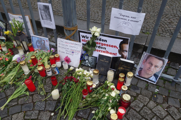 Ein Mahnmal in Berlin, Deutschland, mit Blumen, Kerzen, Fotos und Plakaten, die vor einem Zaun auf dem Boden plaziert sind.