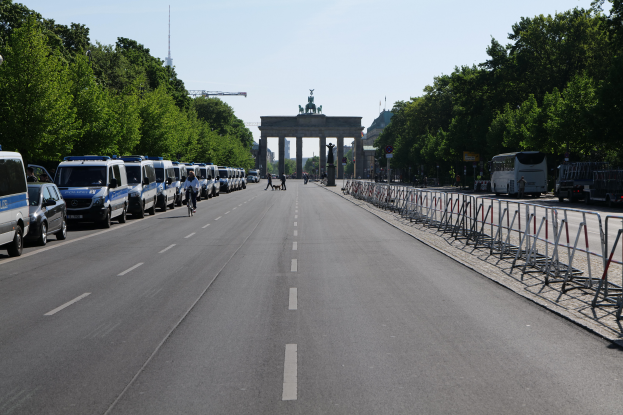 Eine Reihe von Polizeiautos, die auf einer Straße vor dem Brandenburgertor in Berlin, Deutschland, geparkt sind, mit Menschen auf Fahrrädern und Stehenden, Barrieren, Bäumen und einem Bogen mit Statuen im Hintergrund.