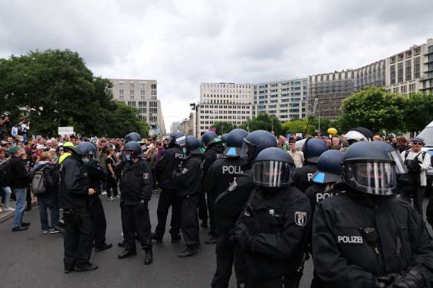 Große Gruppe von Polizisten vor einer Menge auf einer baumbestandenen Straße in Berlin, einige Demonstranten halten Kameras unter einem bewölkten Himmel.