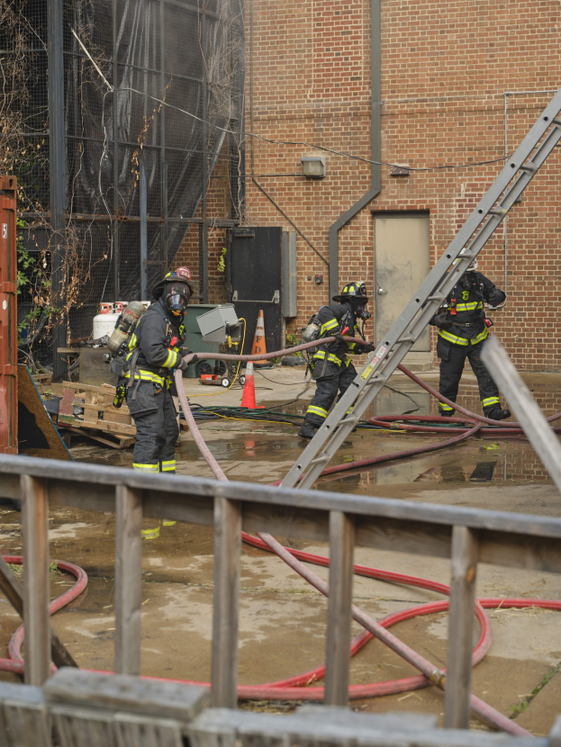 Feuerwehrleute in Helmen arbeiten daran, ein Gebäude Feuer zu löschen, umgeben von Ausrüstung und einem Metallzaun, mit einem Baum und Himmel im Hintergrund.