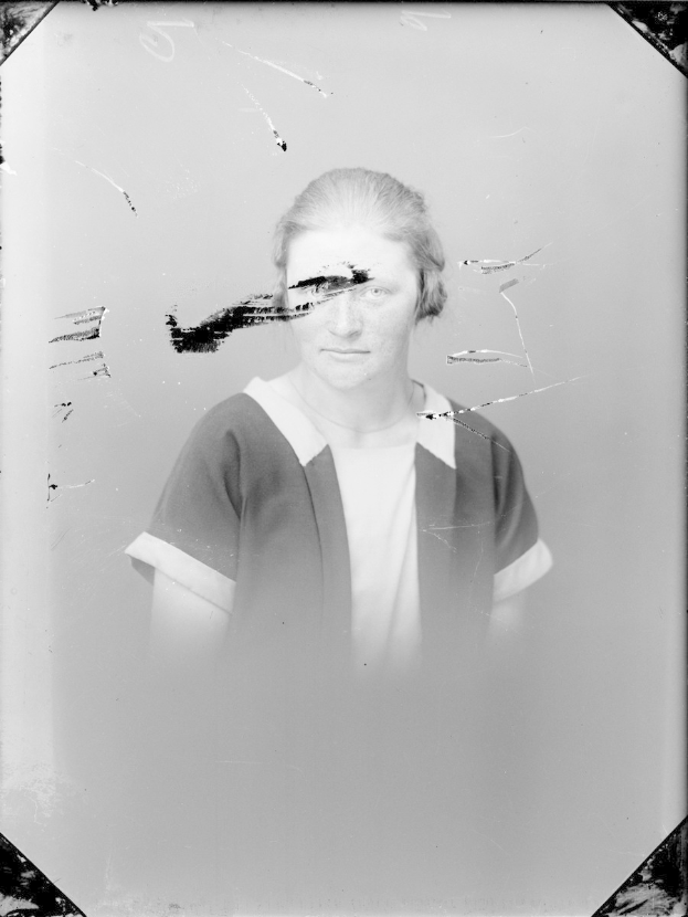 Schwarz-weiß-Foto einer Frau mit einem zerbrochenen Glasfenster im Hintergrund, ihr Gesicht ist deutlich zu erkennen, mit direktem Augenkontakt und leicht geöffneten Lippen, das Haar ist zurückgekämmt, die Hände sind vor der Brust verschränkt.