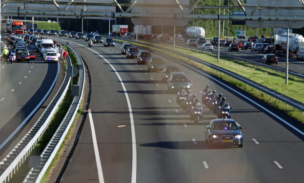 Autobahn mit dichtem Verkehr aus Autos und Motorrädern, gesäumt von Geländern und Gras, mit Bäumen, Pfählen, Schildern und einer Brücke mit Metallstangen im Hintergrund.