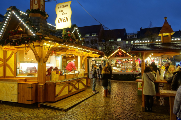 Ein belebter Weihnachtsmarkt auf einer Kopfsteinpflasterstraße bei Nacht, mit Menschen um dekorierte Stände, Gebäuden im Hintergrund, Bäumen und einem klaren Himmel.