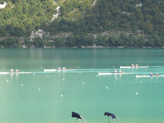 Eine Gruppe von Menschen bei einem Ruderwettbewerb auf einem Gewässer mit Bäumen und einem Hügel im Hintergrund, mit Kleidung über Ständen am Boden.