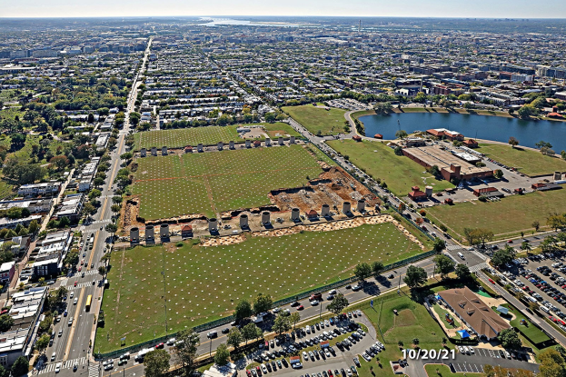 Luftbild einer Stadt mit Gebäuden, Bäumen, Gras, Straßen mit Fahrzeugen, Wasser und Wolken, das ein großes zentrales Feld zeigt, das für einen Park vorgesehen ist.