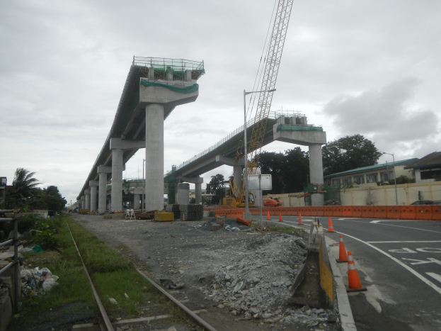 Baustelle mit einer Brücke im Hintergrund, Straße mit Absperrgittern, Bahnschiene, Steine, Gras, Bäume, Gebäude und bewölkter Himmel.
