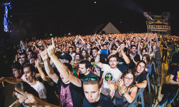 Eine große Menschenmenge auf einem Musikfestival mit Menschen, die ihre Hände heben, einige sitzen und andere stehen, in der Nähe eines Metallzauns mit Gebäuden und Lichtern im Hintergrund unter einem dunklen Himmel.
