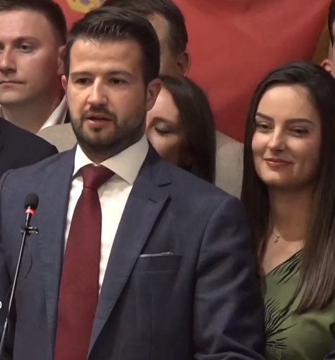 Ein Mann im Anzug spricht in ein Mikrofon an einem Rednerpult, mit einem Banner dahinter und einer Menge davor.