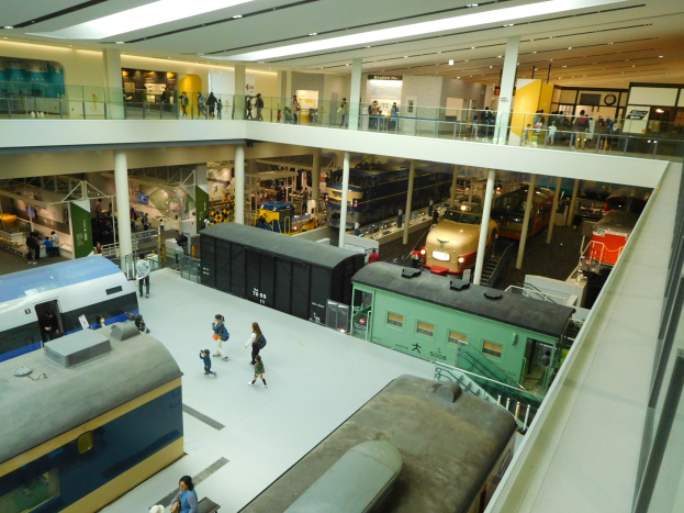 Eine großzügige Museumsausstellung mit verschiedenen Arten von Zügen, Menschen, Geländern, Säulen, Deckenleuchten, Informationsschildern und anderen verwandten Gegenständen.