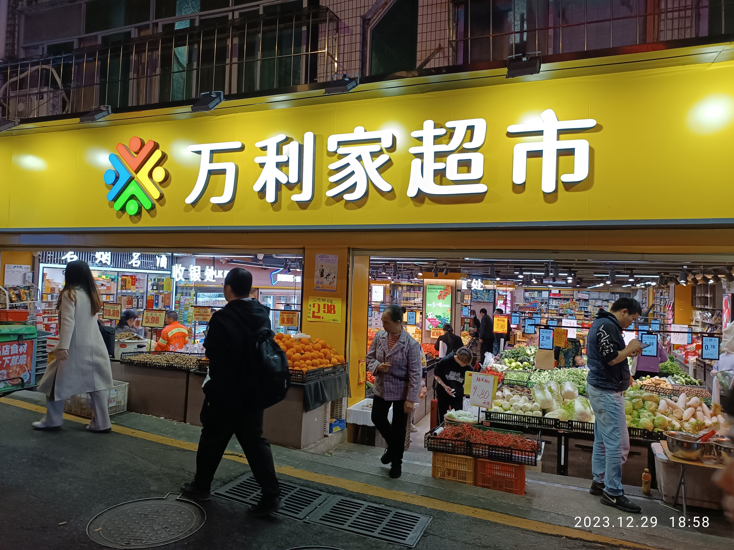 Eine Gruppe von Menschen geht an einem chinesischen Supermarkt vorbei mit sichtbaren Produkträumen, Informationsschildern und Beleuchtung, vor städtischen Gebäuden und einem Gullideckel auf der Straße.