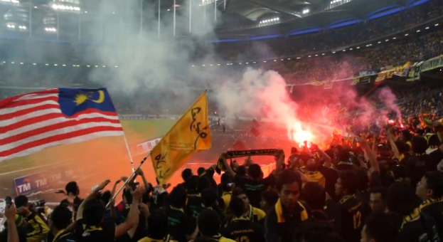 Eine große Menschenmenge in einem Stadion mit Bengalos, Fahnen und Rauch, mit Schildern, die "Malaysia vs Malaysia Live Streaming" anzeigen.