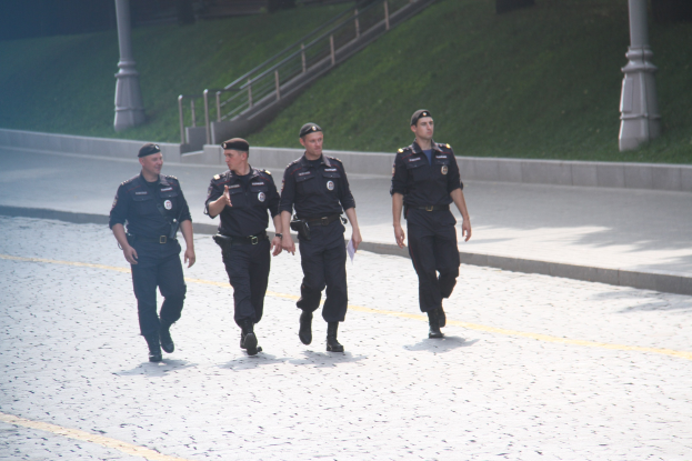 Drei Polizeibeamte in Uniform und Mützen gehen eine Straße mit Pfählen, Geländern, Treppen und Gras im Hintergrund entlang.