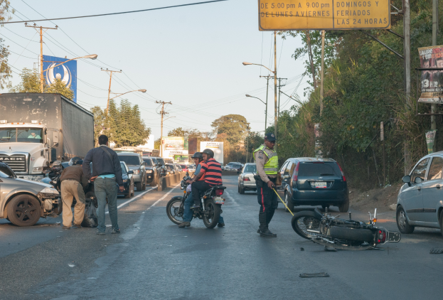 Eine Gruppe von Menschen umringt ein verunglücktes Motorrad am Straßenrand mit mehreren Fahrzeugen, darunter ein Lastwagen, und einem Hintergrund aus Bäumen, Pfosten, Lampen und Schildern unter dem Himmel.
