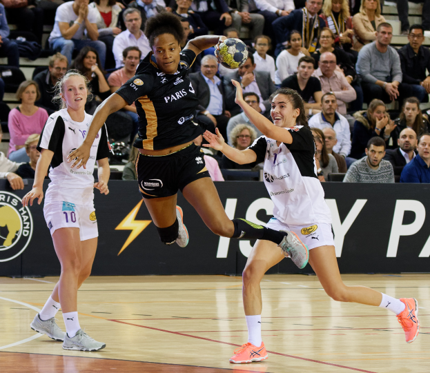 Eine Gruppe von Frauen beim Handballspielen auf einem Platz, mit einem Ball in der Mitte der Aktion, während Zuschauer im Hintergrund sitzen und ein Banner mit Text und Logos sichtbar ist.