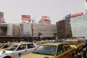 Eine Stadtansicht mit Fahrzeugen auf der Straße, Gebäuden, Bäumen, Masten mit Schildern und Text auf den Schildern, einem Zug im Hintergrund und einem Geländer unten rechts.