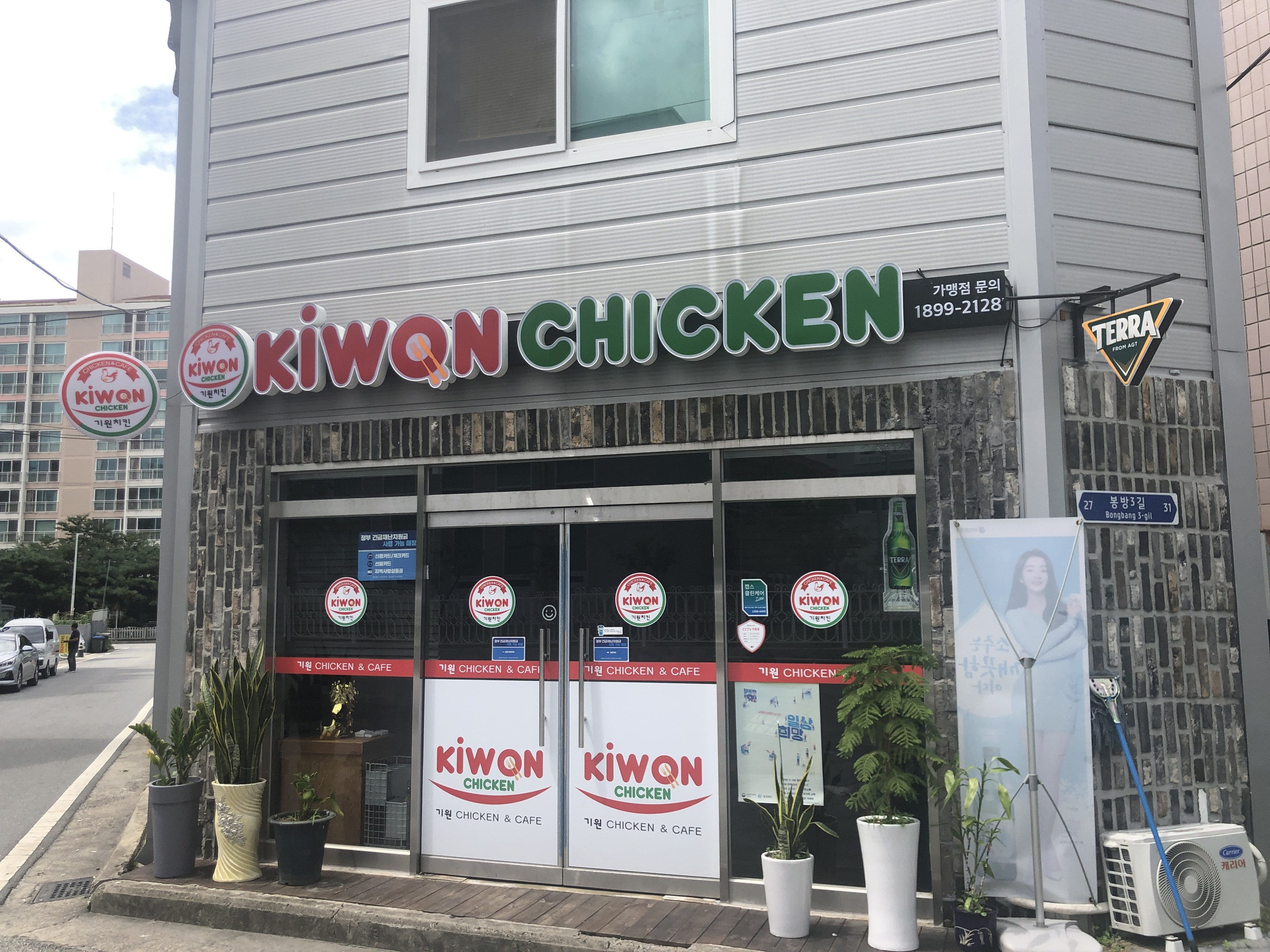 Außenansicht eines Kiwon Chicken-Restaurants in Seoul, Südkorea, mit Glasfenstern und -türen, Topfpflanzen, einem Banner, einer Klimaanlage, einem Pfosten, Kabeln, Bäumen und Fahrzeugen auf der Straße unter einem bewölkten Himmel.