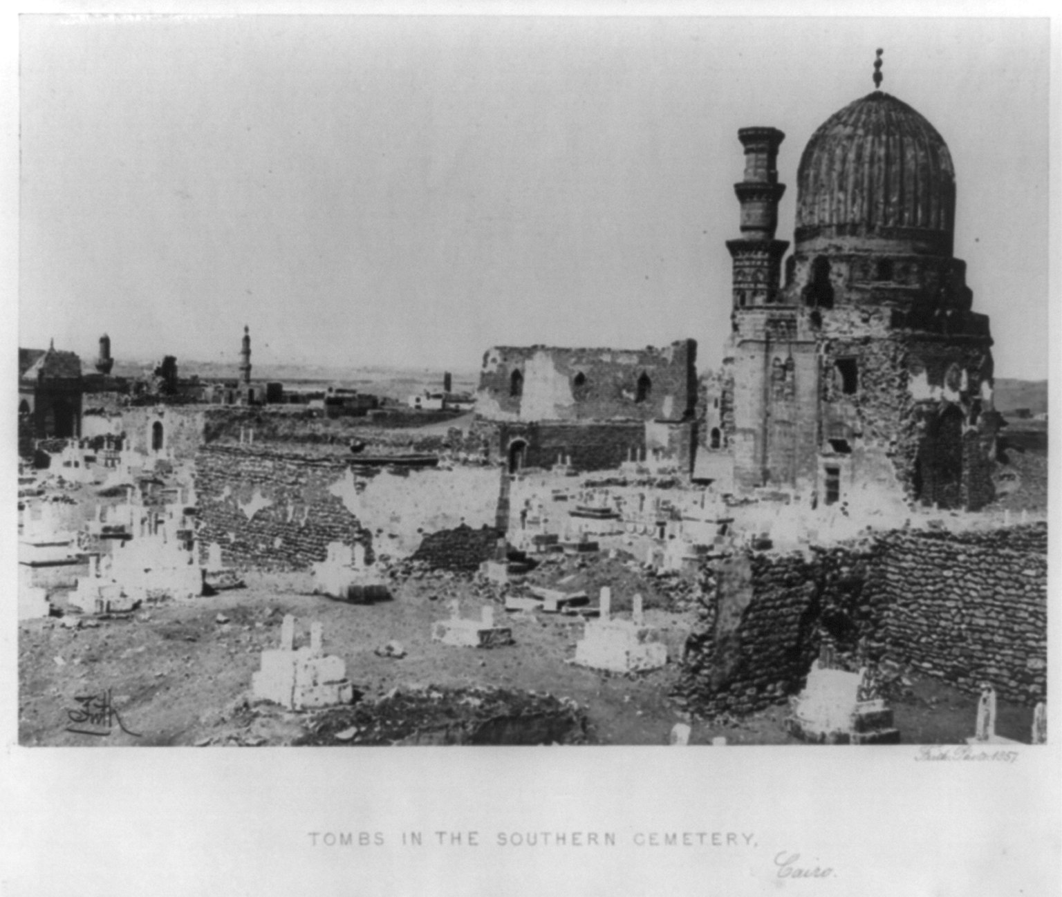 Ein Schwarz-Weiß-Foto eines Friedhofs mit zahlreichen Grabsteinen, einer Moschee im Hintergrund und Text am unteren Bildrand.