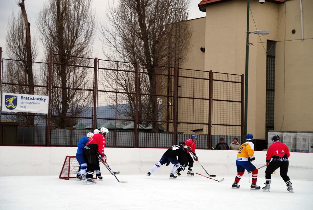 Menschen, die Eis-hoc-key auf einer Eisbahn mit Gebäuden, Bäumen, einer Straßenlaterne, einer Namensschilder und Zäunen im Hintergrund unter einem klaren Himmel spielen.