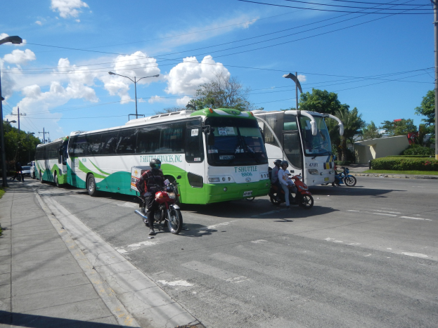 Ein grüner und weißer Shuttlebus, der auf der Straße geparkt ist, mit Motorradfahrern davor, einem grasbewachsenen Fußweg links und Gebäuden, Bäumen, Laternenpfählen und einem klaren blauen Himmel im Hintergrund.