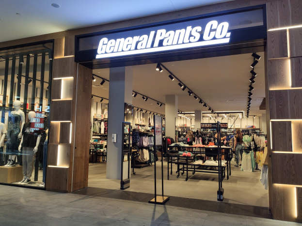 Innenansicht eines General Pants Co. Geschäfts in einem Einkaufszentrum, mit Schaufensterpuppen in Kleidern, hängenden Kleidungsstücken, Tischen mit Gegenständen, Säulen, einer wandmontierten Namensschild und Deckenbeleuchtung.