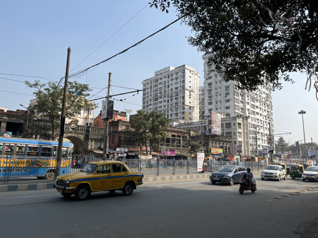 Gelbes Taxi auf einer belebten Straße in Mumbai fahren, mit hohen Gebäuden, Bäumen, Strommasten und Schildern im Hintergrund unter einem sichtbaren Himmel.
