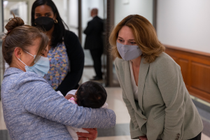 Senatorin Amy Klobuchar, mit Mund-Nasen-Schutz, hält ein Baby, während sie mit einer Gruppe von Menschen drinnen steht, wobei eine Tür, eine Wand und Deckenleuchten im Hintergrund zu sehen sind.