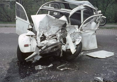 Schwer beschädigtes Frontende eines Wagens, der auf der Seite der Straße mit Bäumen und einem Gebäude im Hintergrund gestrandet ist.