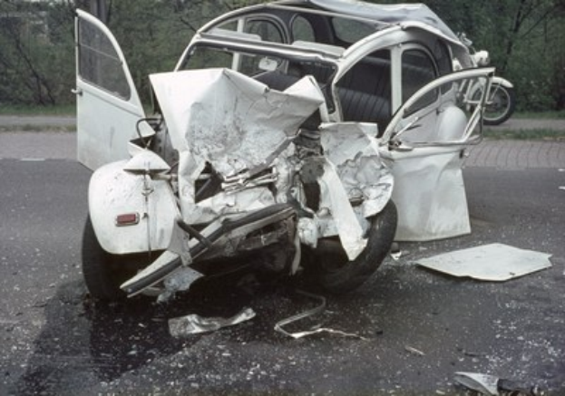 Schwer beschädigtes Frontende eines Wagens, der auf der Seite der Straße mit Bäumen und einem Gebäude im Hintergrund gestrandet ist.