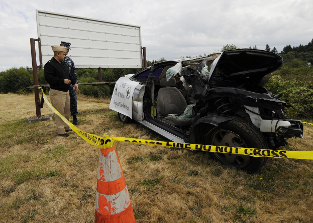 Ein Polizeibeamter in Uniform steht neben einem schwer beschädigten Auto am Straßenrand mit eingedrütkter Haube und zersplitterten Fenstern, bei einer Verkehrskegel und eine mit einem Band markierte Stange, vor einem Hintergrund von Bäumen und einem bewölktem Himmel.