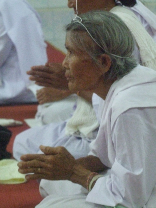 Alte Frau im Krankenhausbett liegend mit gefalteten Händen, geschlossenen Augen und Kopf auf den Händen ruhend, umgeben von Menschen im Hintergrund.