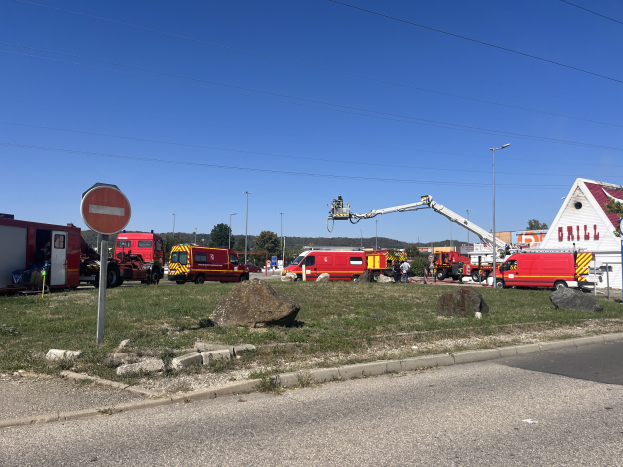 Feuerwehrl├Ąrmen auf der Seite einer Stra├če in der N├Ąhe einer Tankstelle geparkt, mit umgebender Gras, Steinen, einem Schild, einem Haus, Masten, einem Kran, Dr├Ąhten, B├Ąumen und einem bew├Âlktem Himmel.